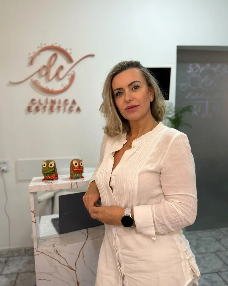 Dina Costa de blusa branca na recepção da clínica, com o letreiro dc Clínica Estética na parede